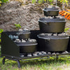 Stacked black cast iron Dutch ovens with charcoal briquettes on a sturdy steel outdoor cooking table, set on grass with a stone wall and gloves in the background, ideal for farm, garden, camping, and best sellers collections.