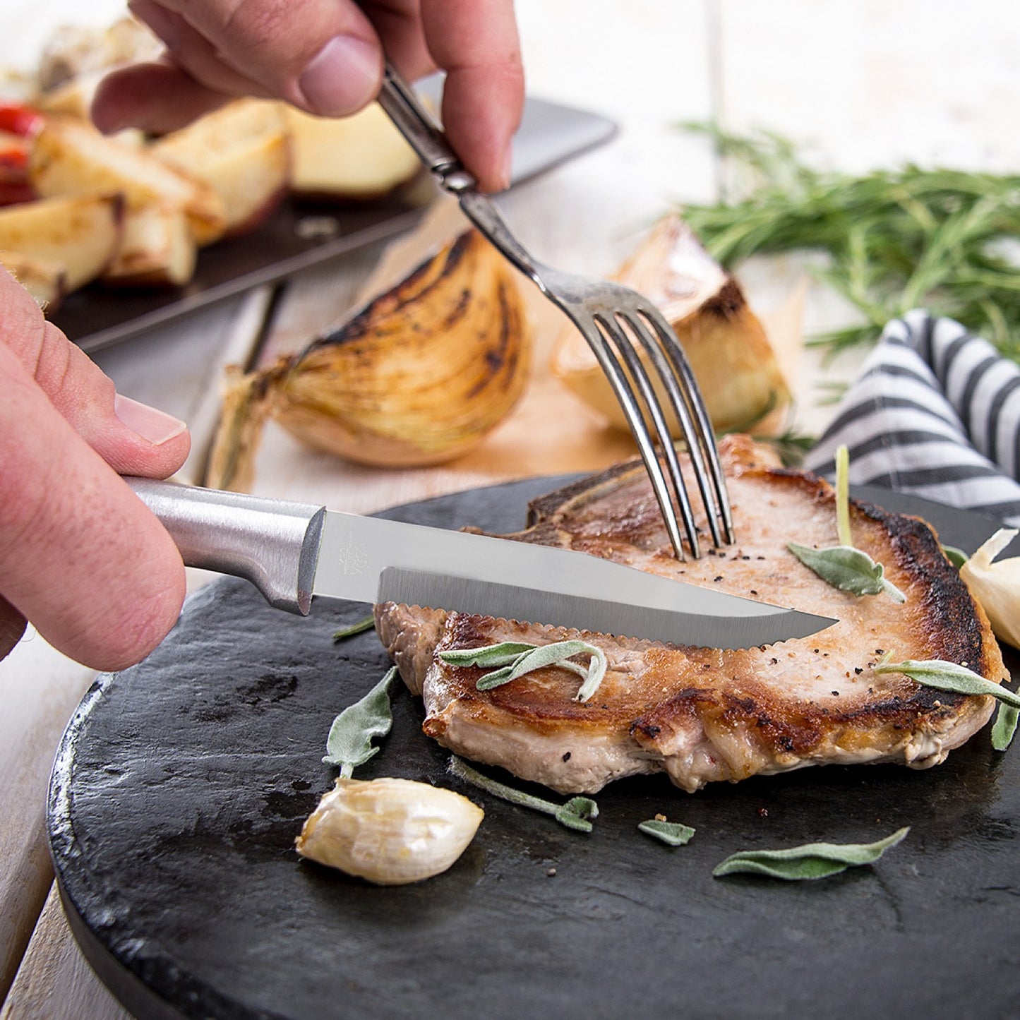 Handcrafted serrated steak knife with high carbon surgical stainless steel blade and solid aluminum handle slicing a grilled pork chop garnished with herbs and garlic, ideal for Farm, Garden & Tool, Kitchen & Food, Knives & Axes, and USA Made collections.
