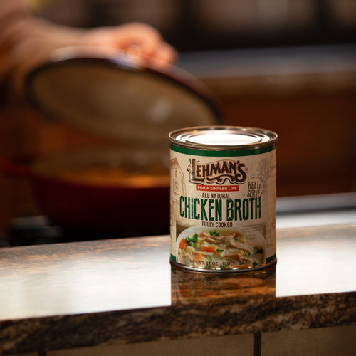 27 oz can of Lehman's all natural fully cooked chicken broth placed on a kitchen counter with a person lifting a pot lid in the background, ideal for Farm, Kitchen & Food, Emergency Supplies, and Best Sellers collections.