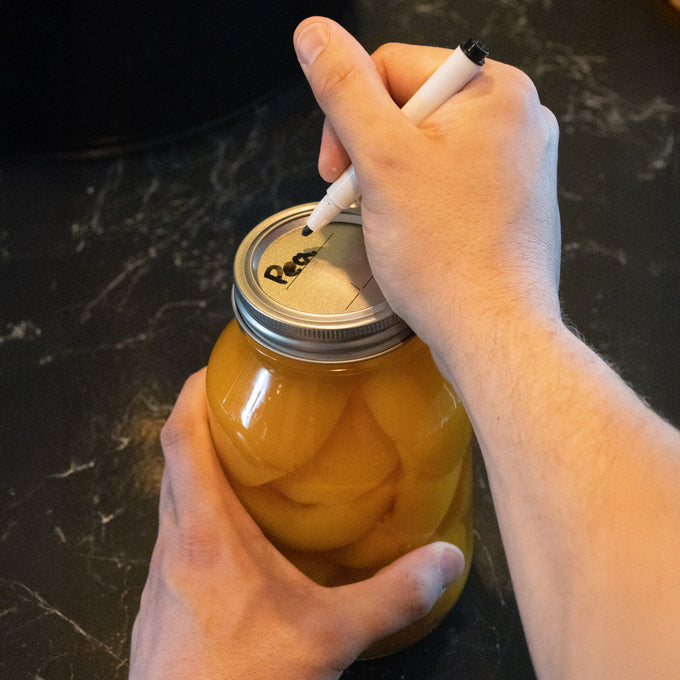 Person writing a label on a silver-colored regular-mouth canning jar lid sealing a jar filled with preserved peaches, highlighting food preservation in Kitchen & Food and Canning collections.