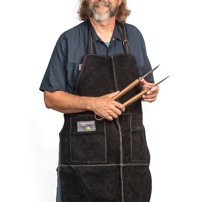Man wearing an adjustable black leather grill apron holding wooden-handled barbecue tongs, suitable for Kitchen & Food, Grilling, and Kitchen Textiles collections.