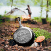 Handcrafted cast iron skillet with a tent and campsite design, seasoned and made in USA, shown outdoors on forest ground near mushrooms, perfect for camping and cookware collections.