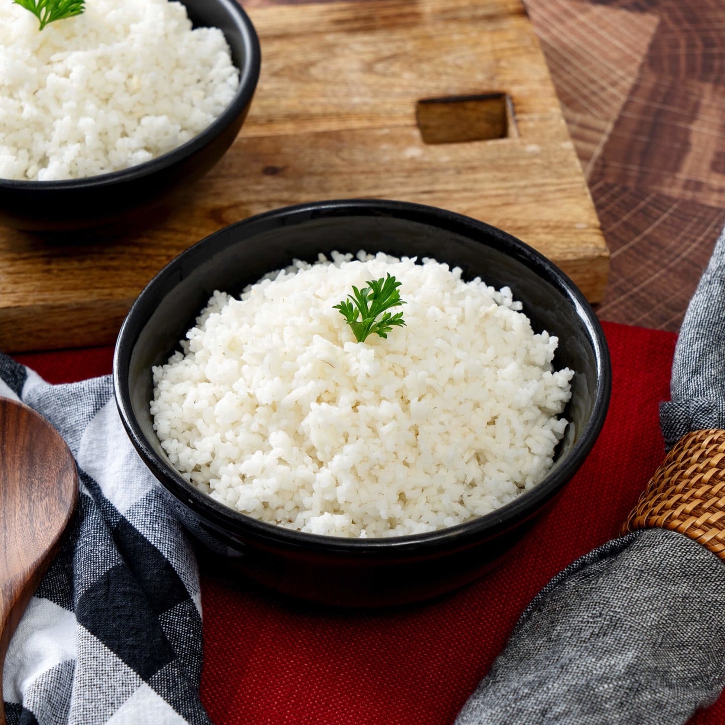 Black bowl filled with fluffy white rice garnished with a small sprig of parsley, set on a red cloth with rustic wooden cutting board and kitchen linens nearby, ideal for emergency supplies and USA Made survival food collections.