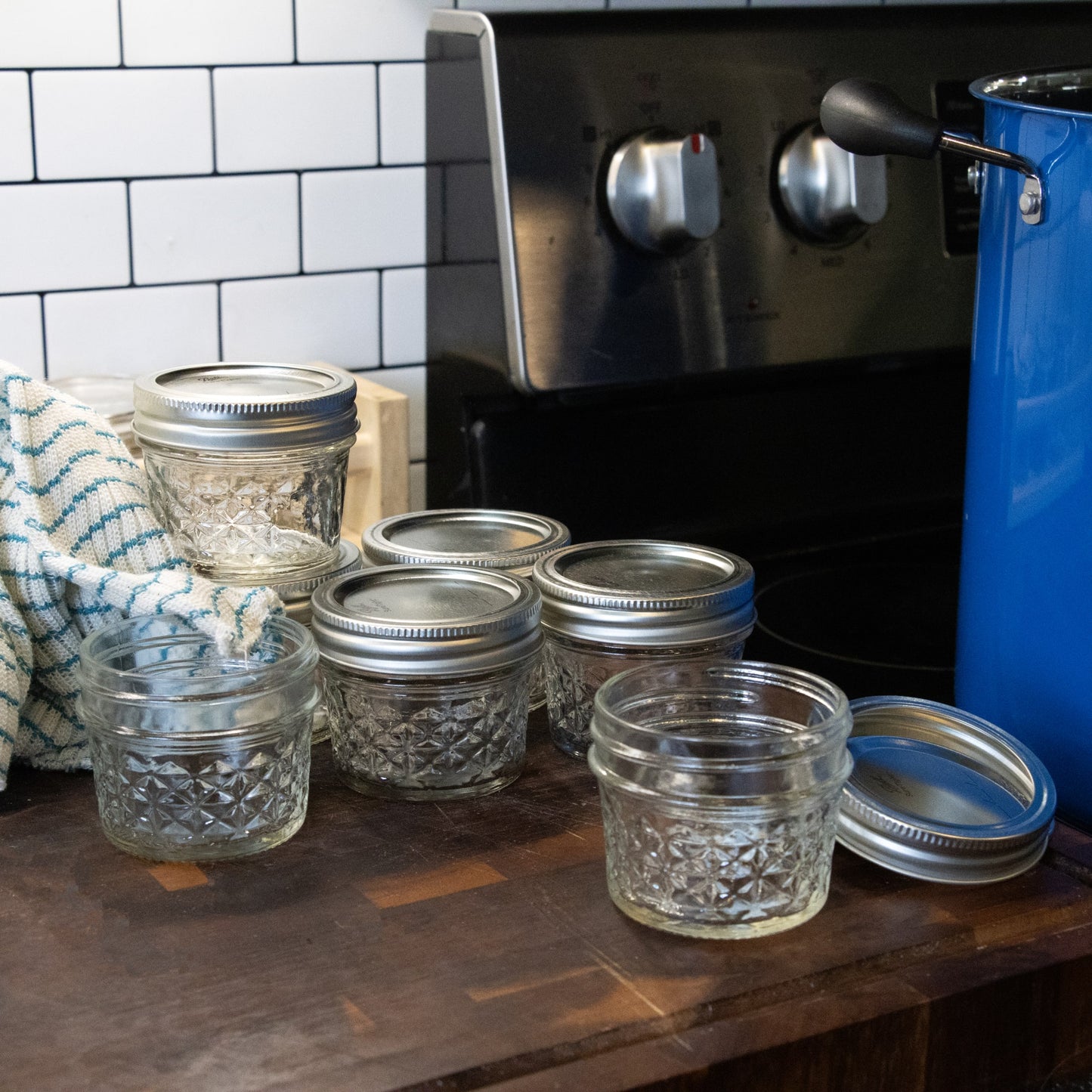 Set of clear quilted glass jars with silver metal lids on a wooden kitchen counter near a stove and blue pot, handcrafted for food preservation and canning, part of USA Made and Kitchen & Food collections.