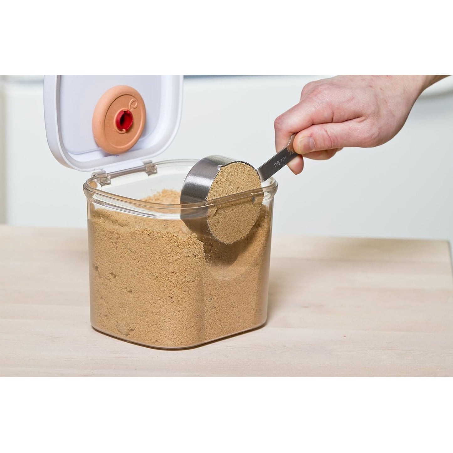 Clear storage container with a snap-shut lid and terra cotta moisture keeper inside, filled with brown sugar, shown with a hand scooping sugar using a metal measuring cup, ideal for Kitchen & Food and Storage collections.