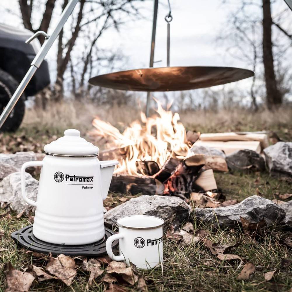 White enamel Petromax coffee pot and matching mug with black logo, placed on a black trivet near a campfire outdoors, perfect for Camping, Kitchen & Food, and Brewing Coffee and Tea collections.