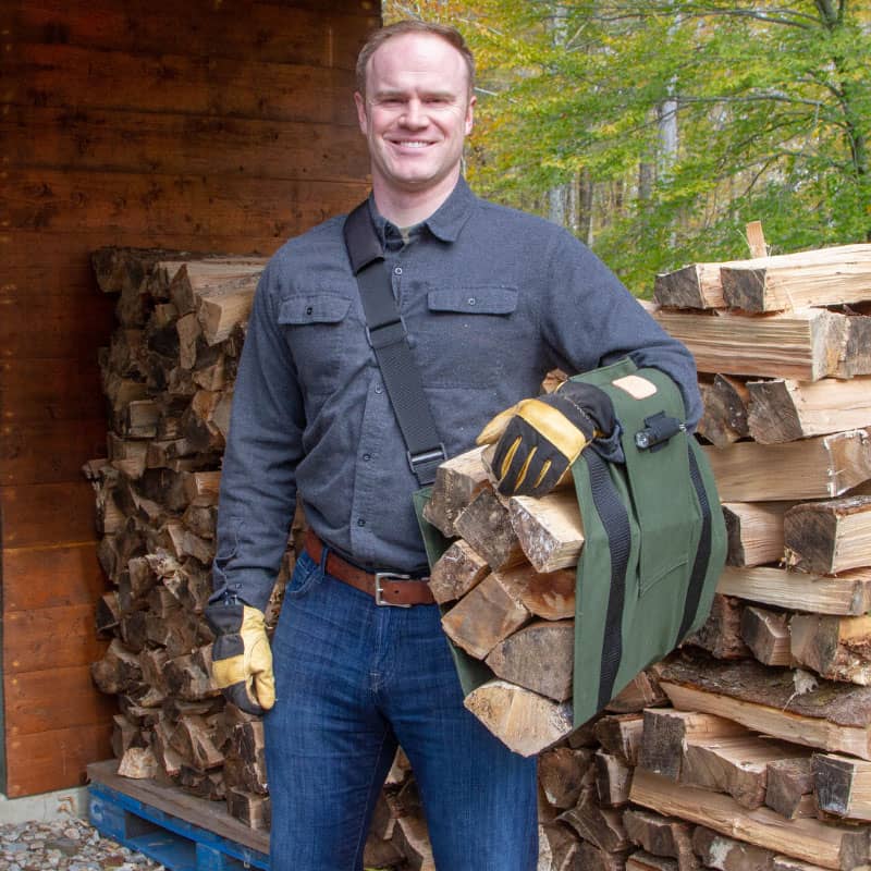 Man wearing gloves and a dark gray shirt carrying split firewood logs in a green sling with adjustable black straps, designed for ergonomic wood hauling, ideal for Farm, Garden & Tool and Wood Cutting and Hauling collections, USA made and best sellers.