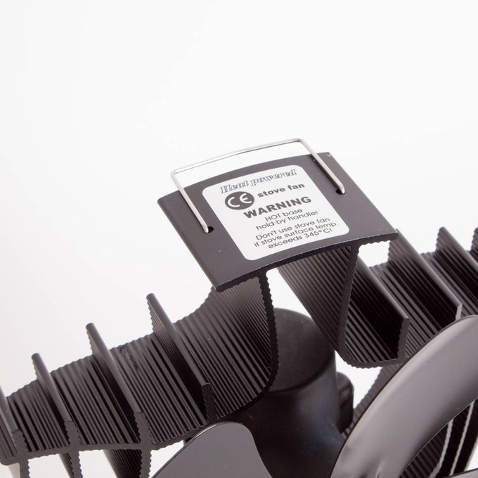 Close-up of a black heat-activated oscillating stove fan with curved finned blades and a warning label, designed to improve warm air circulation in stoves and hearth accessories collections.
