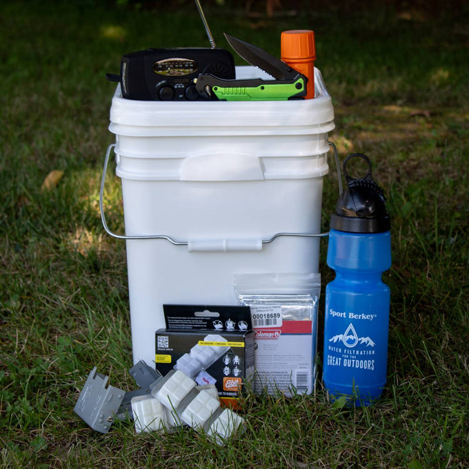 White plastic bucket filled with emergency supplies including a hand crank radio, folding knife, waterproof matches, water filter bottle, fuel tablets, and water purification packets for camping, farm, garden, and emergency use.