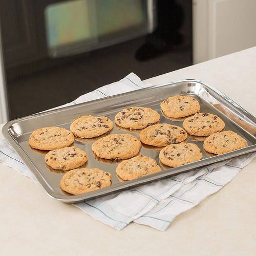 Polished stainless steel cookie sheet holding a batch of freshly baked chocolate chip cookies on a striped kitchen towel, suitable for baking supplies, cookware, and holiday hosting collections.