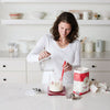 Woman in a bright kitchen mixing sourdough starter ingredients in a glass container with a red silicone spatula, surrounded by baking supplies for Kitchen & Food and Baking Supplies collections.