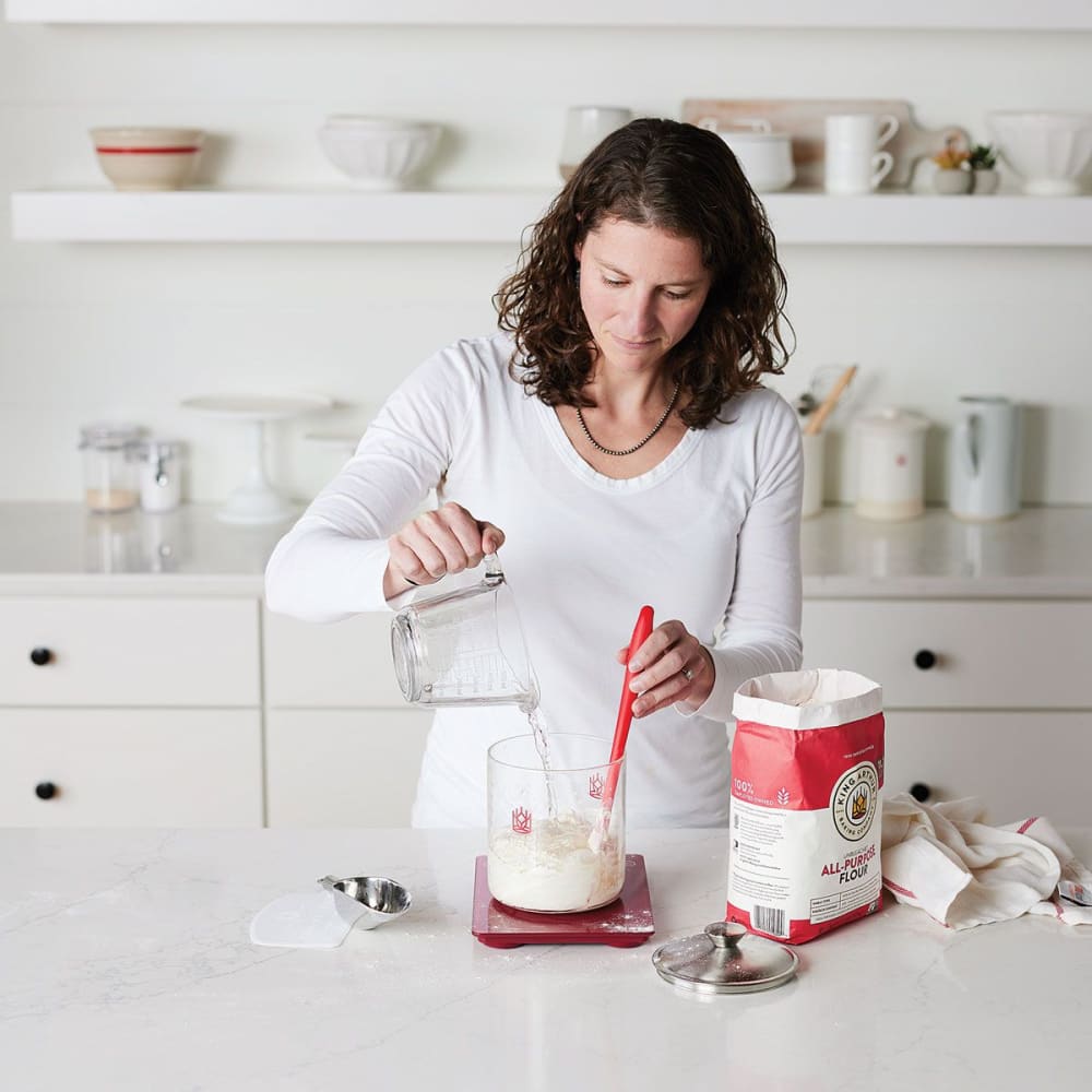 Woman in a bright kitchen mixing sourdough starter ingredients in a glass container with a red silicone spatula, surrounded by baking supplies for Kitchen & Food and Baking Supplies collections.