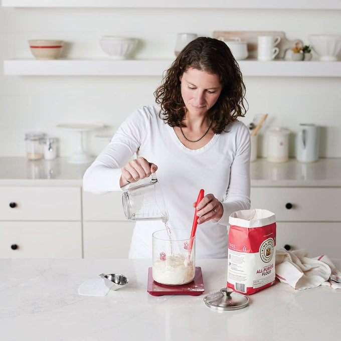 Woman in a bright kitchen mixing sourdough starter ingredients in a glass container with a red silicone spatula, surrounded by baking supplies for Kitchen & Food and Baking Supplies collections.