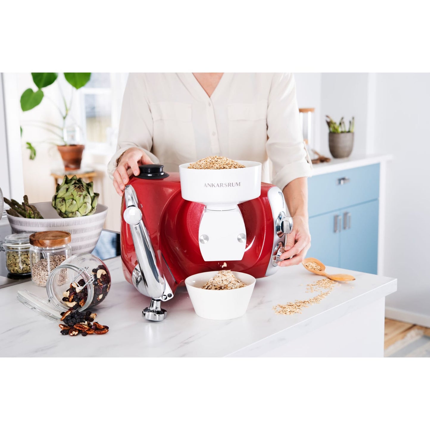 Person using a red grain flake mill attachment on a kitchen mixer to freshly roll oats with scattered grains and a bowl of rolled oats on a white countertop in a bright kitchen setting ideal for Kitchen & Food, Baking Supplies, and Grain Mills collections.