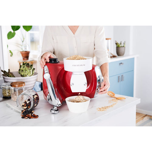 Person using a red grain flake mill attachment on a kitchen mixer to freshly roll oats with scattered grains and a bowl of rolled oats on a white countertop in a bright kitchen setting ideal for Kitchen & Food, Baking Supplies, and Grain Mills collections.