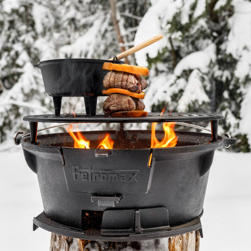 Petromax cast iron fire grill with height-adjustable grate cooking skewered meat and vegetables alongside a pot over a blazing fire in a snowy outdoor setting, ideal for Farm, Garden & Tool, Stoves, Camping, Grilling, and Best Sellers collections.
