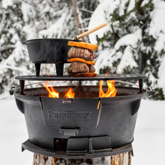 Petromax cast iron fire grill with height-adjustable grate cooking skewered meat and vegetables alongside a pot over a blazing fire in a snowy outdoor setting, ideal for Farm, Garden & Tool, Stoves, Camping, Grilling, and Best Sellers collections.