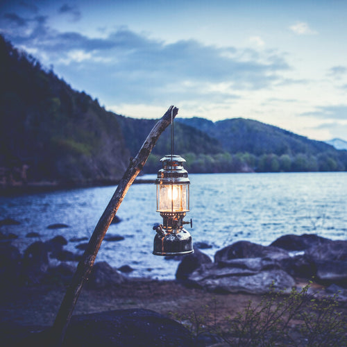 Handcrafted Petromax HK500 kerosene pressure lamp emitting warm light by a lakeside at dusk, ideal for camping, outdoors, farm, garden, and emergency supplies.
