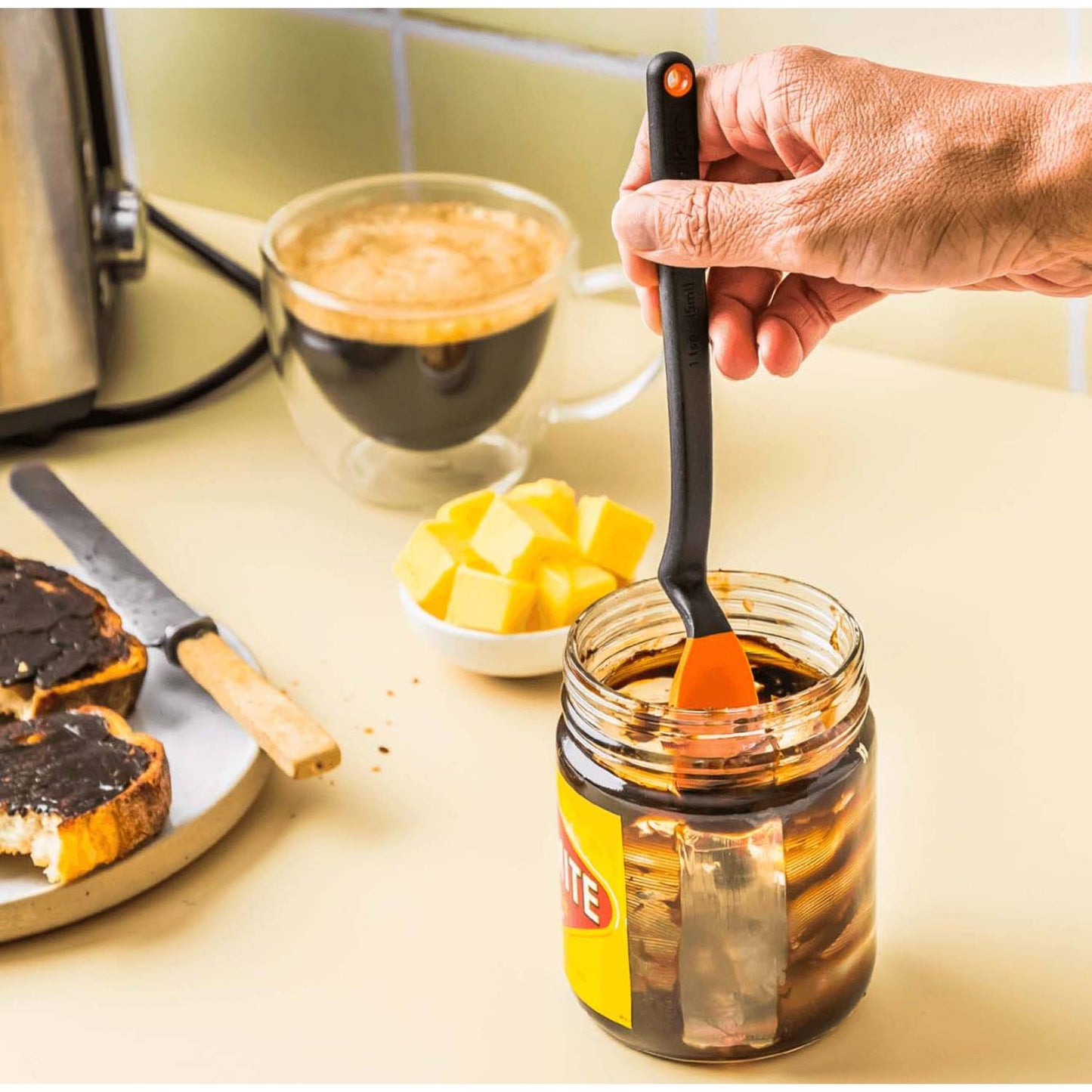 Hand using a flexible orange silicone spoon with a black handle to scoop spread from a jar, shown with toast, butter cubes, and a cup of coffee, in Kitchen & Food and Food Prep collections.