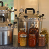 Three handcrafted large stainless steel pressure canners with secure locking lids and pressure gauges on a kitchen counter, surrounded by glass jars of preserved vegetables and sauces, fitting Kitchen & Food, Food Preservation, Canning, Pressure Canners, USA Made, and Best Sellers collections.