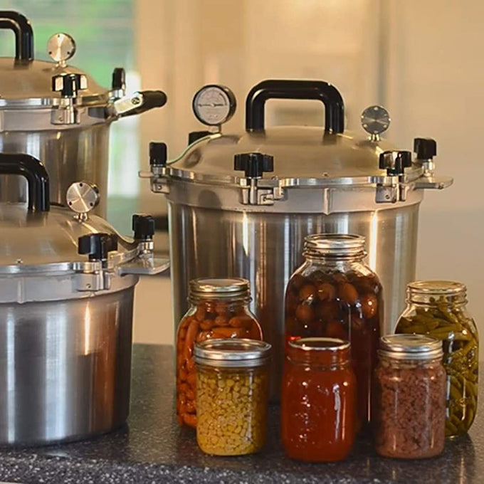 Three handcrafted large stainless steel pressure canners with secure locking lids and pressure gauges on a kitchen counter, surrounded by glass jars of preserved vegetables and sauces, fitting Kitchen & Food, Food Preservation, Canning, Pressure Canners, USA Made, and Best Sellers collections.