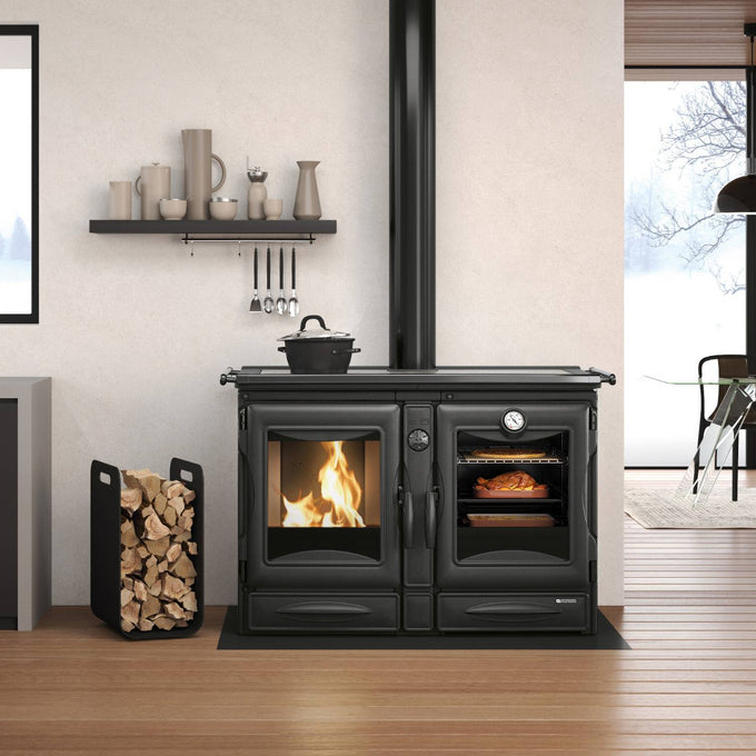 Black enameled cast iron wood cook stove with visible fire and oven cooking bread, featured in a modern kitchen setting with firewood stored nearby, part of Stoves and Cookstoves collections.