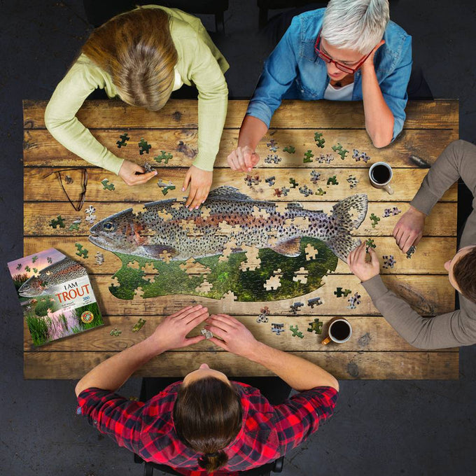 Four people gathered around a wooden table assembling a uniquely shaped trout puzzle with 300 pieces, accompanied by a trout facts booklet and coffee mugs, in a cozy game setting.