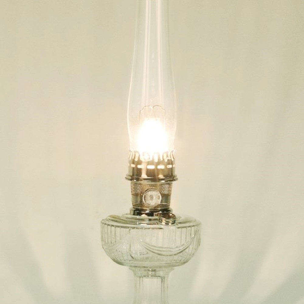 Lit vintage-style clear glass oil lamp with a tall chimney and metal burner, featured in Lamps & Lighting, Parts and Accessories, Wicks and Mantles, Lamp Burners and Collars, Best Sellers, and Promo Eligible collections.