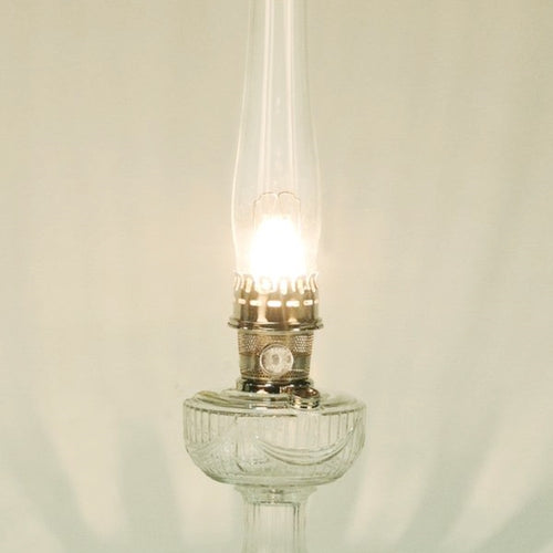 Lit vintage-style clear glass oil lamp with a tall chimney and metal burner, featured in Lamps & Lighting, Parts and Accessories, Wicks and Mantles, Lamp Burners and Collars, Best Sellers, and Promo Eligible collections.