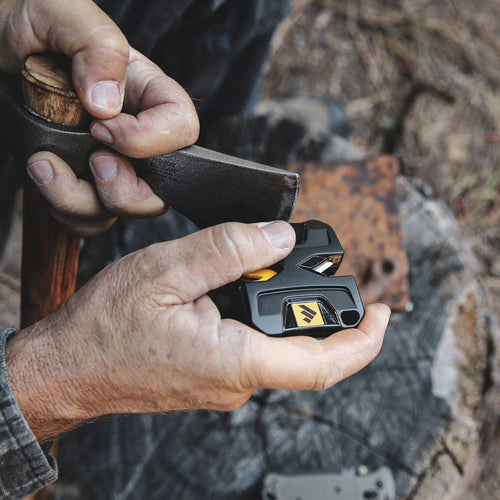 Hands holding a compact black and yellow portable sharpener, sharpening the blade of a small hand axe outdoors on a tree stump, suitable for farm, garden, and tool use.