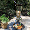 Outdoor scene featuring a shiny metal camping stove with cooking attachment, a plate of breakfast with eggs, bacon, and beans, fresh herbs in a paper bag, all set on a wooden table in a woodland setting, ideal for Farm, Garden & Tool and Camping collections.