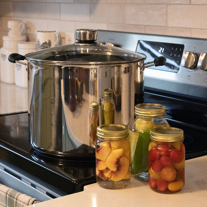 Polished stainless steel 21.5-quart water bath canner with glass lid and black handles, displayed on a stove next to three sealed jars of preserved fruits in a kitchen environment.