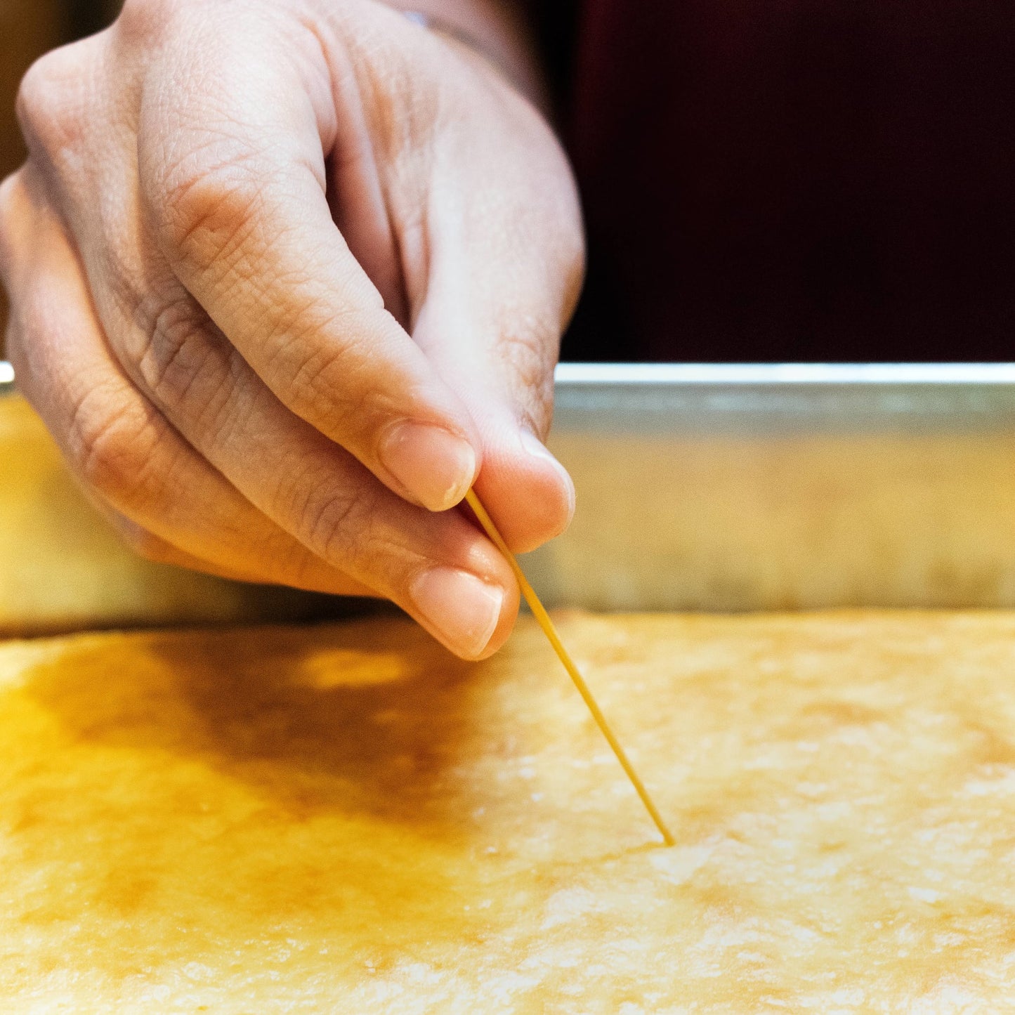 Close-up of hand using a slender straw from a handcrafted Amish-made broom cake tester to check doneness of a baked cake in a metal pan, part of Kitchen & Food and Kitchen Gadgets collections.