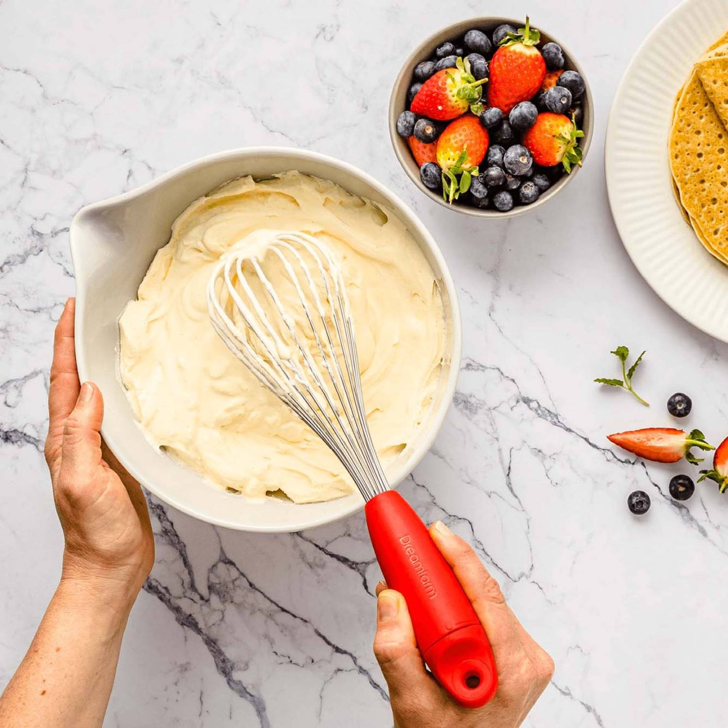 Hands holding a red-handled whisk mixing creamy batter in a white mixing bowl on a marble countertop, with fresh strawberries, blueberries, and crackers nearby, ideal for Kitchen & Food, Kitchen Gadgets, Cooking and Baking Helpers, Best Sellers, Promo Eligible collections.