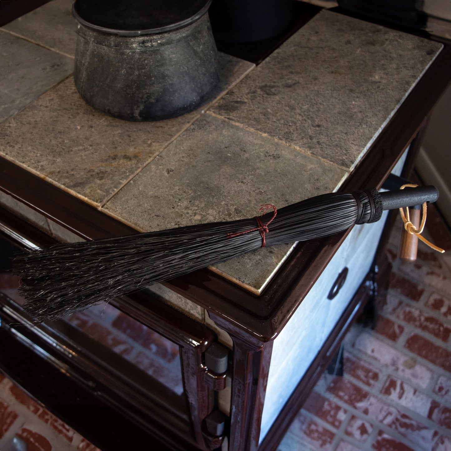 Handmade Amish small hand broom with tightly bundled natural fibers and black wooden handle, resting on a rustic tiled stove surface, ideal for hearth cleaning in USA Made and Hearth Accessories collections.