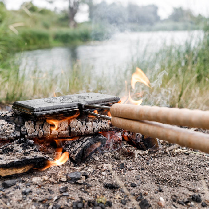 Cast iron waffle iron with wooden handles cooking over an open campfire near a river, ideal for outdoor grilling and included in Farm, Garden & Tool, Camping, and Best Sellers collections.