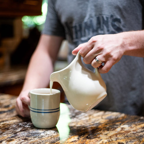 Handcrafted cream-colored stoneware pitcher pouring liquid into a ceramic mug with blue stripes, ideal for Kitchen & Food, Dinnerware, Storage and Serving, USA Made, and Best Sellers collections.