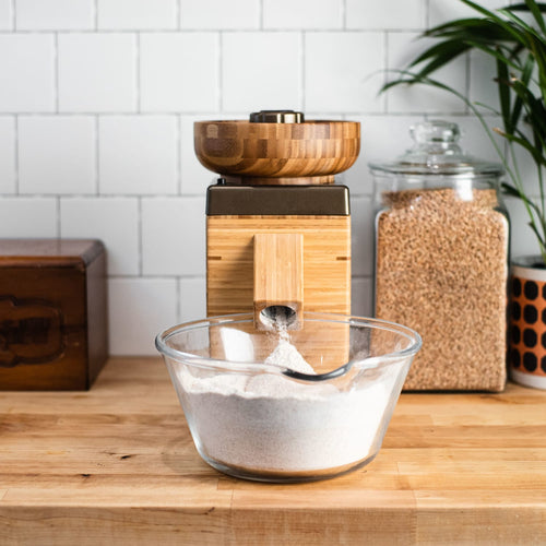 Electric grain grinder with a natural bamboo finish dispensing freshly milled flour into a clear glass bowl, pictured on a wooden countertop with grains in a glass jar nearby, ideal for Kitchen & Food, Grain Mills, Best Sellers, and Bread Making Supplies collections.