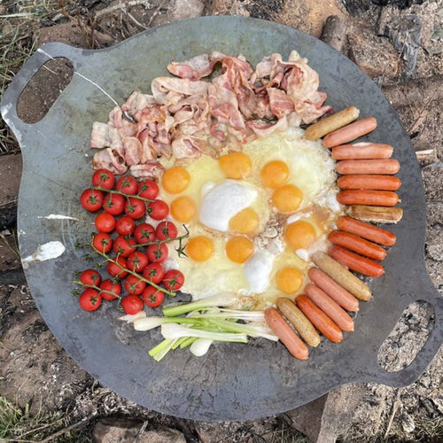 Large steel griddle with dual handles cooking a colorful breakfast of fried eggs, cherry tomatoes, bacon, sausages, and green onions over an open campfire, ideal for Farm, Garden & Tool, Camping, Best Sellers, and Promo Eligible collections.
