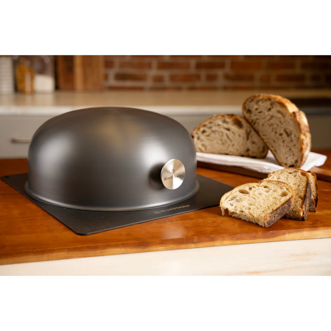 Matte black baking shell with a metal knob, placed on a flat baking sheet beside sliced artisan bread, ideal for Kitchen & Food, Cookware, Baking Supplies, and Bread Making collections.