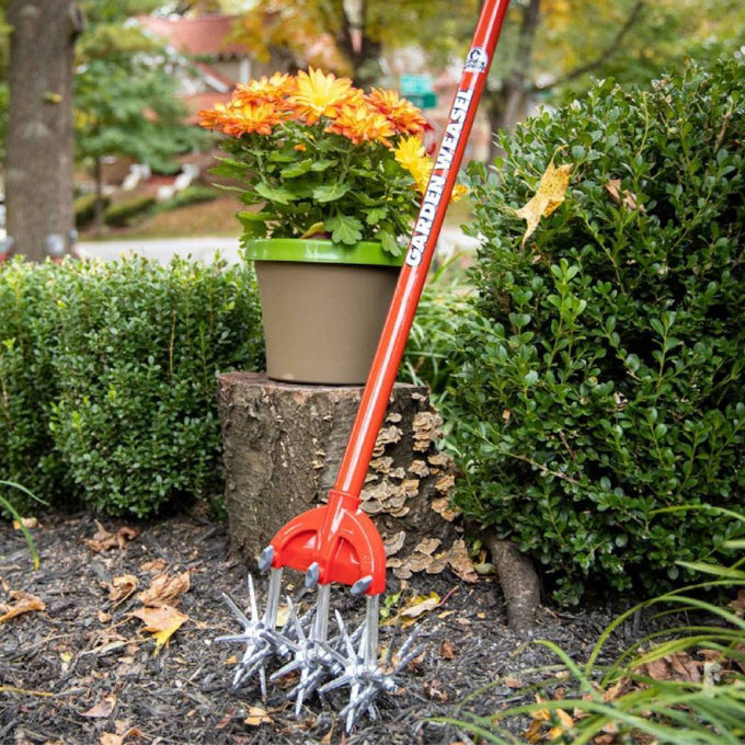 Original Garden Weasel with rust-proof serrated tines and red comfort-grip handle leaning against a tree stump next to potted flowers and green shrubs in a garden setting, featured in Farm, Garden & Tool and Digging and Cultivating collections.