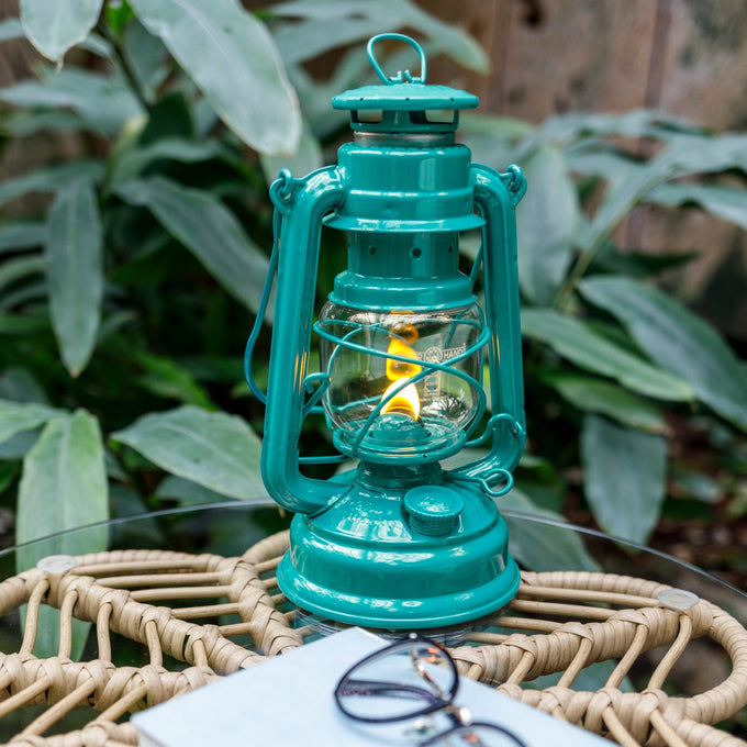 Handcrafted teal galvanized oil lantern with durable thermal heat-resistant globe glowing warmly on a glass table with glasses and notebook nearby, ideal for Lamps & Lighting, Camping, and Emergency Supplies collections.