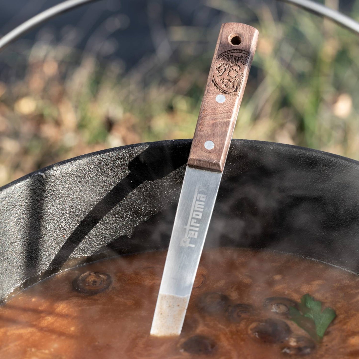 Handcrafted stainless steel soup ladle with engraved wooden handle resting in steaming cast iron pot, designed for generous serving in Farm, Garden & Tool, Camping, and Kitchen & Food collections.
