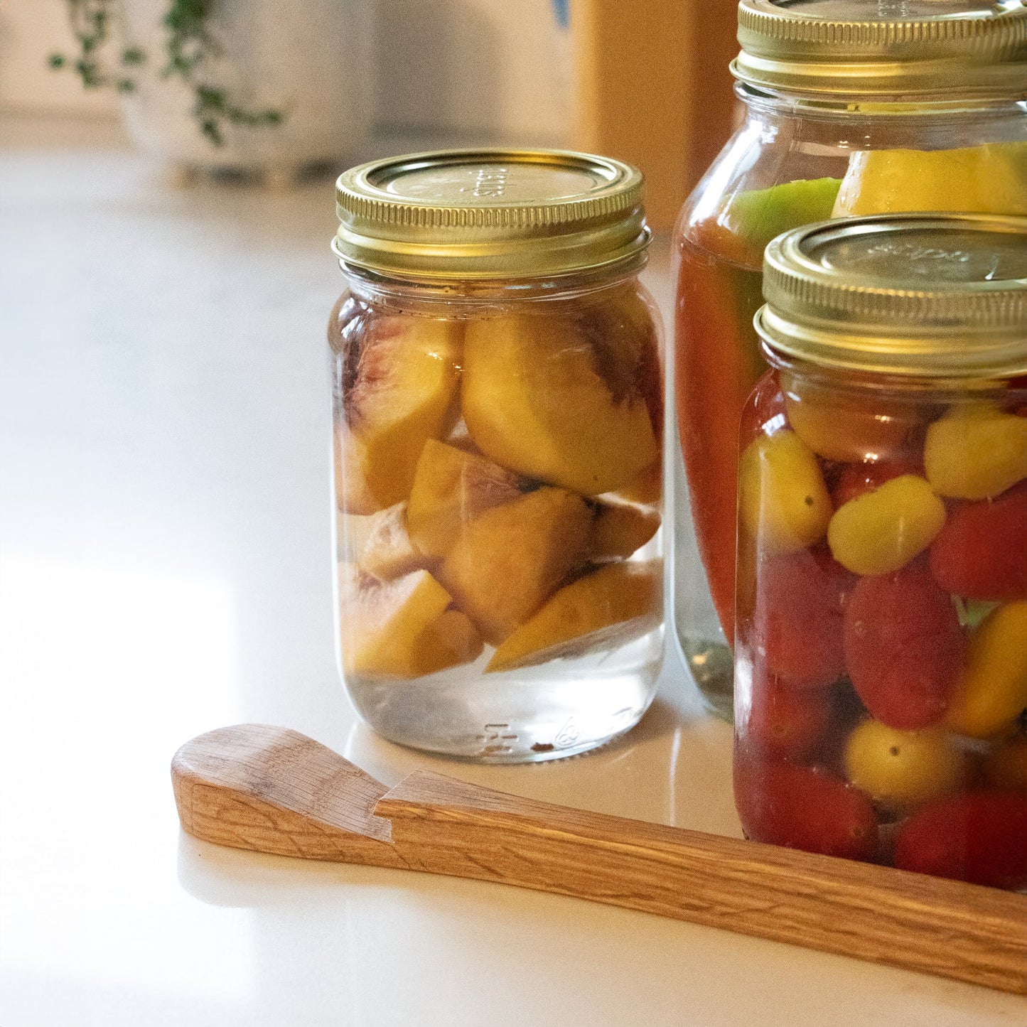 Handcrafted oak jar opener resting beside three sealed mason jars filled with preserved fruit and vegetables, highlighting food preservation and canning accessories collections.