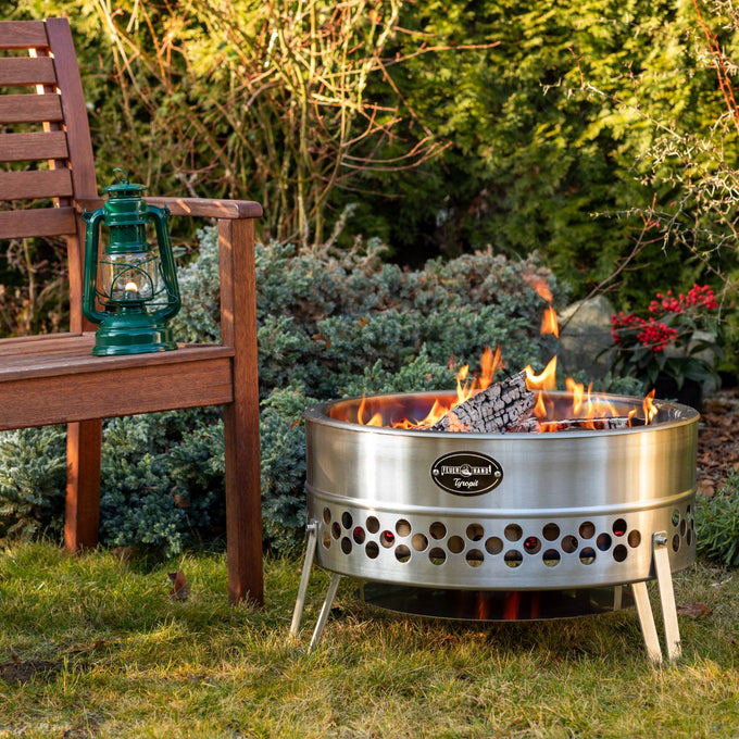 Stainless steel fire bowl with ventilated double-wall design and burning firewood, placed on grass near a wooden bench holding a green lantern, suitable for camping and outdoor use.