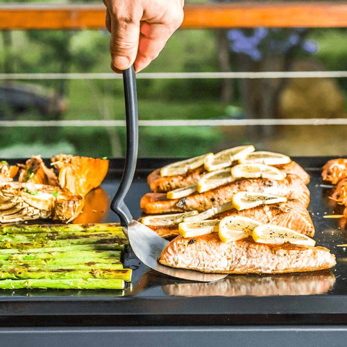Large flexible stainless steel grill spatula with black handle lifting seasoned salmon fillet topped with lemon slices, shown with asparagus and grilled vegetables on flat top grill in outdoor cooking setting