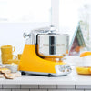 Yellow and silver stand mixer with stainless steel bowl on white kitchen countertop surrounded by ceramic cups, bread slices, and fresh juicer, ideal for Kitchen & Food and Baking Supplies collections.