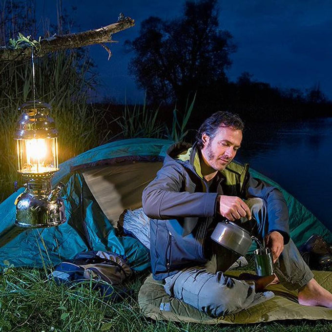 Man camping outdoors at night next to a bright Petromax HK500 kerosene pressure lamp hanging from a tree branch, preparing a hot drink by a tent in a natural setting.