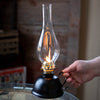 Handcrafted vintage-style oil lamp with black steel font and curved handle, lit flame under clear glass chimney, ideal for Farm, Garden & Tool and Emergency Supplies collections.