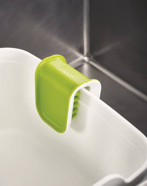 Green and white knife cleaning brush with bristles clamped over the edge of a white basin inside a stainless steel sink, featured in kitchen accessories and cleaning utensils collections.